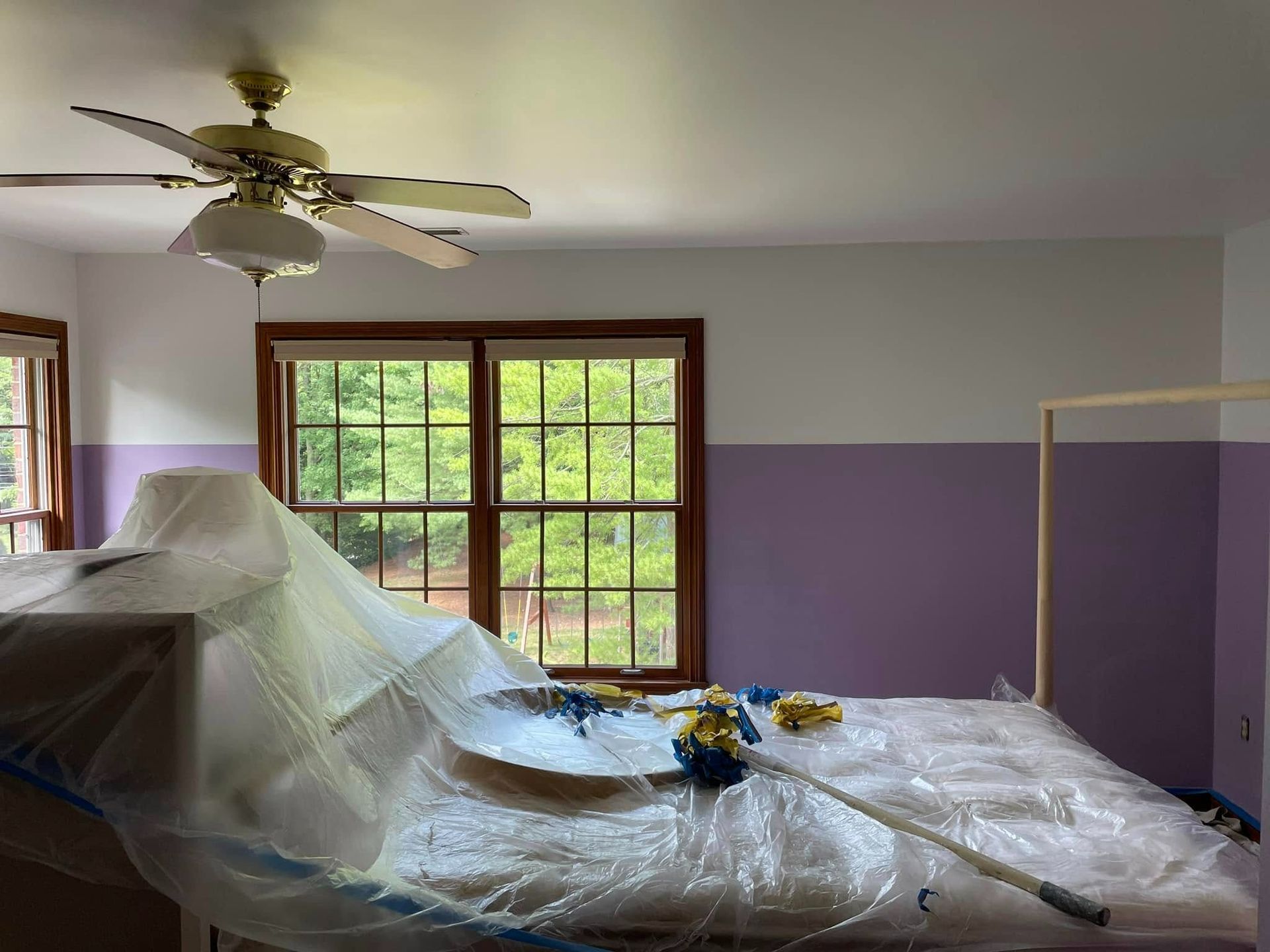 Bedroom being painted; walls are half purple, half white; a bed is covered in plastic, a window in the background.
