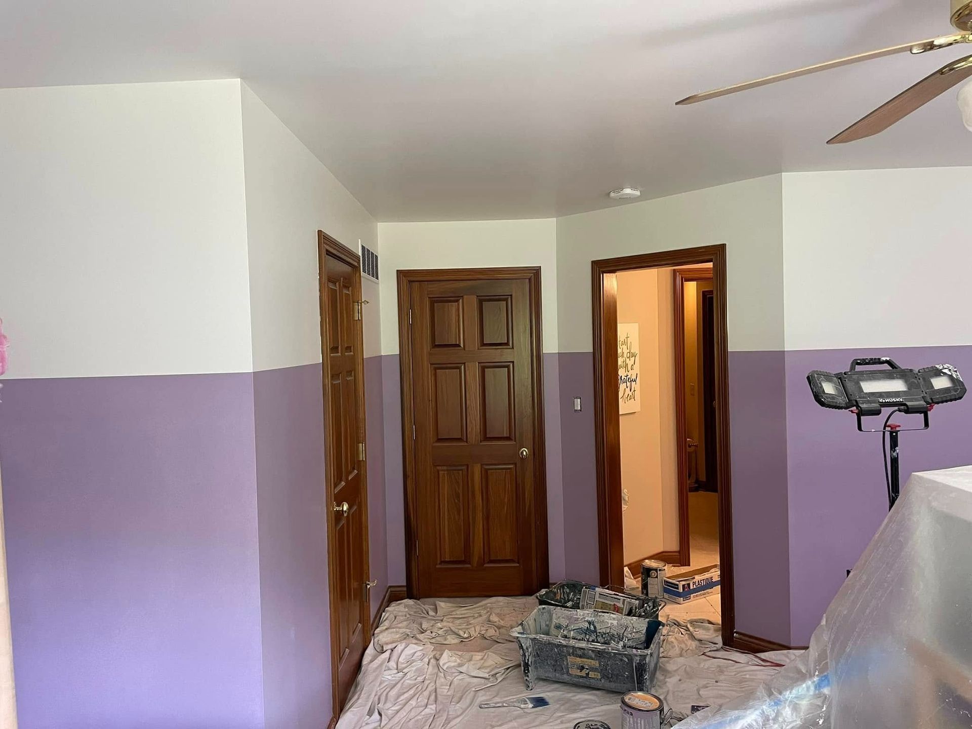 A room with purple and white painted walls; two brown doors; paint supplies on floor.