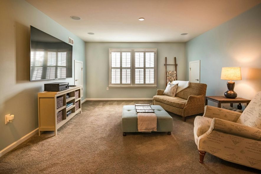 A cozy living room with light blue walls, carpet, a television on the wall, a sofa, a patterned armchair, and an ottoman.