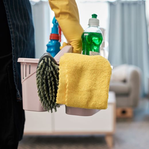 A person wearing a yellow glove carries a light pink caddy filled with a scrub brush, yellow cloth, and green dish soap.