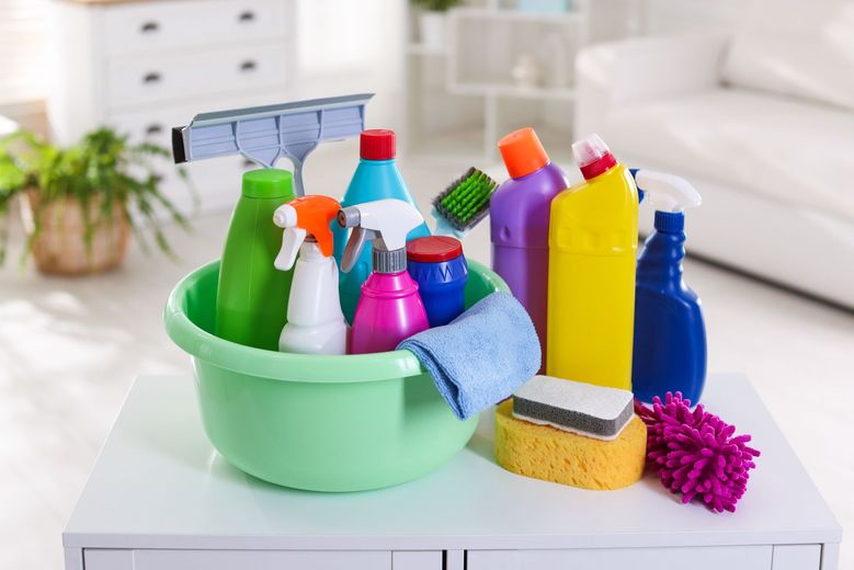 A green basin filled with various colorful cleaning bottles, sponges, and a squeegee sitting on a white table.