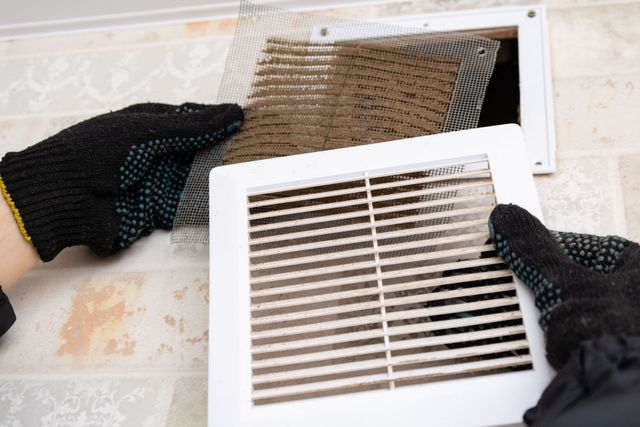 Gloved hands remove a dirty HVAC vent cover with dust and debris on a wall opening