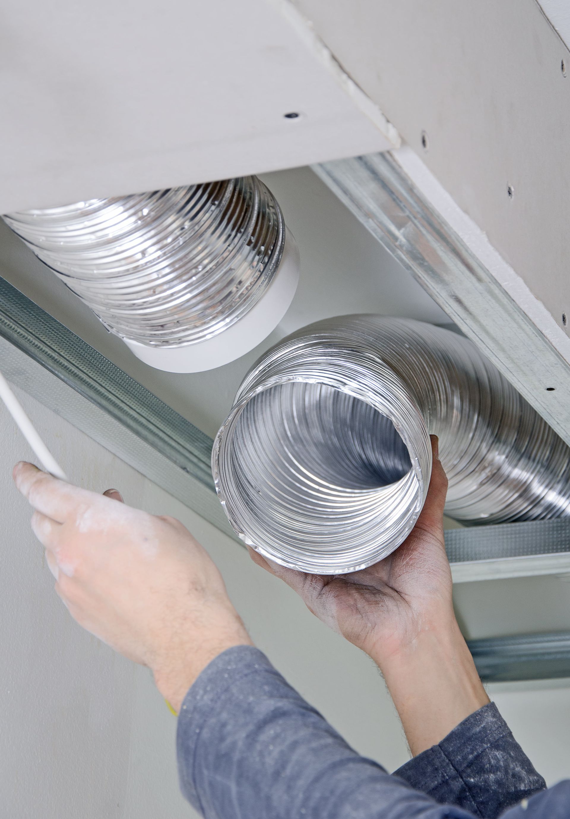 Hands connecting silver flexible HVAC ducts in a ceiling space.