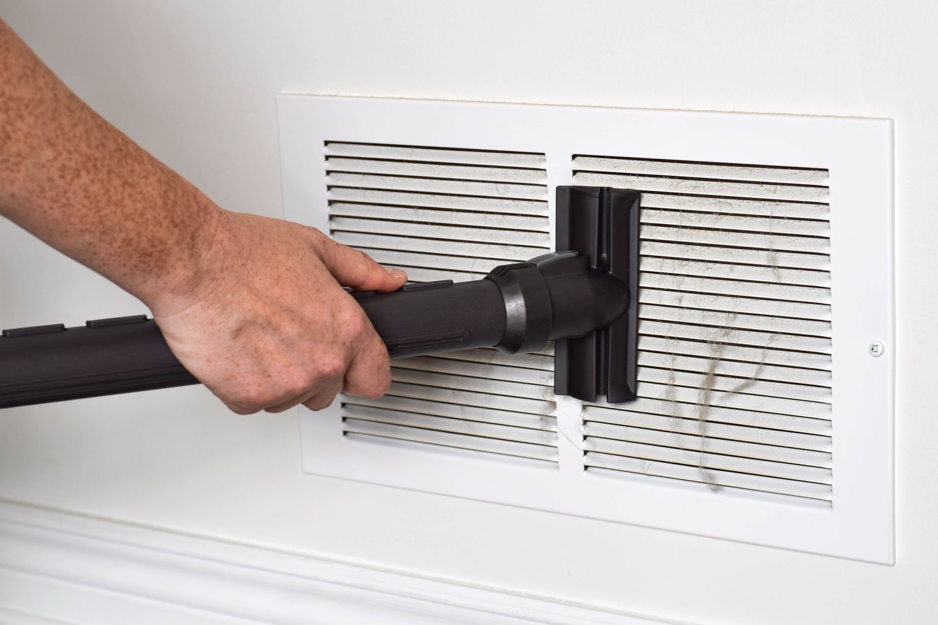 Hand using a vacuum hose to clean a white wall vent cover