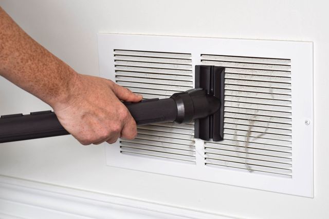 Hand using a vacuum hose to clean a white wall vent cover