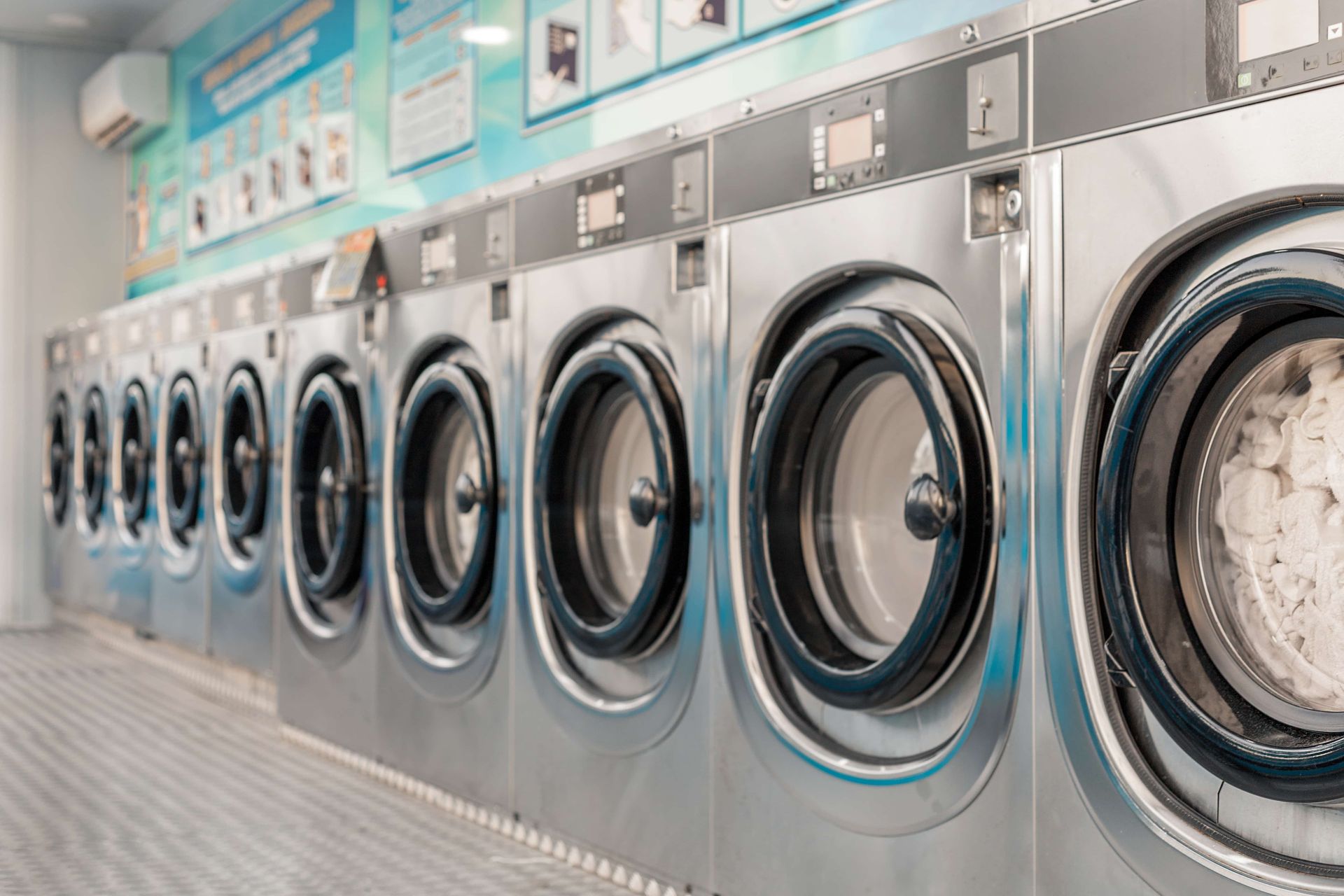 Row of stainless steel washing machines in a laundromat, with one front loader full of laundry