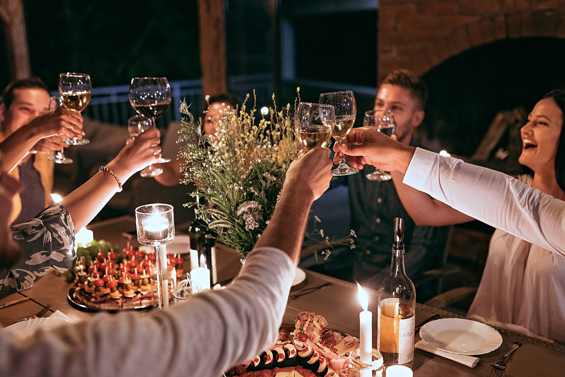 A group of friends laughing and toasting with wine glasses around a dinner table at night, illuminated by candlelight.