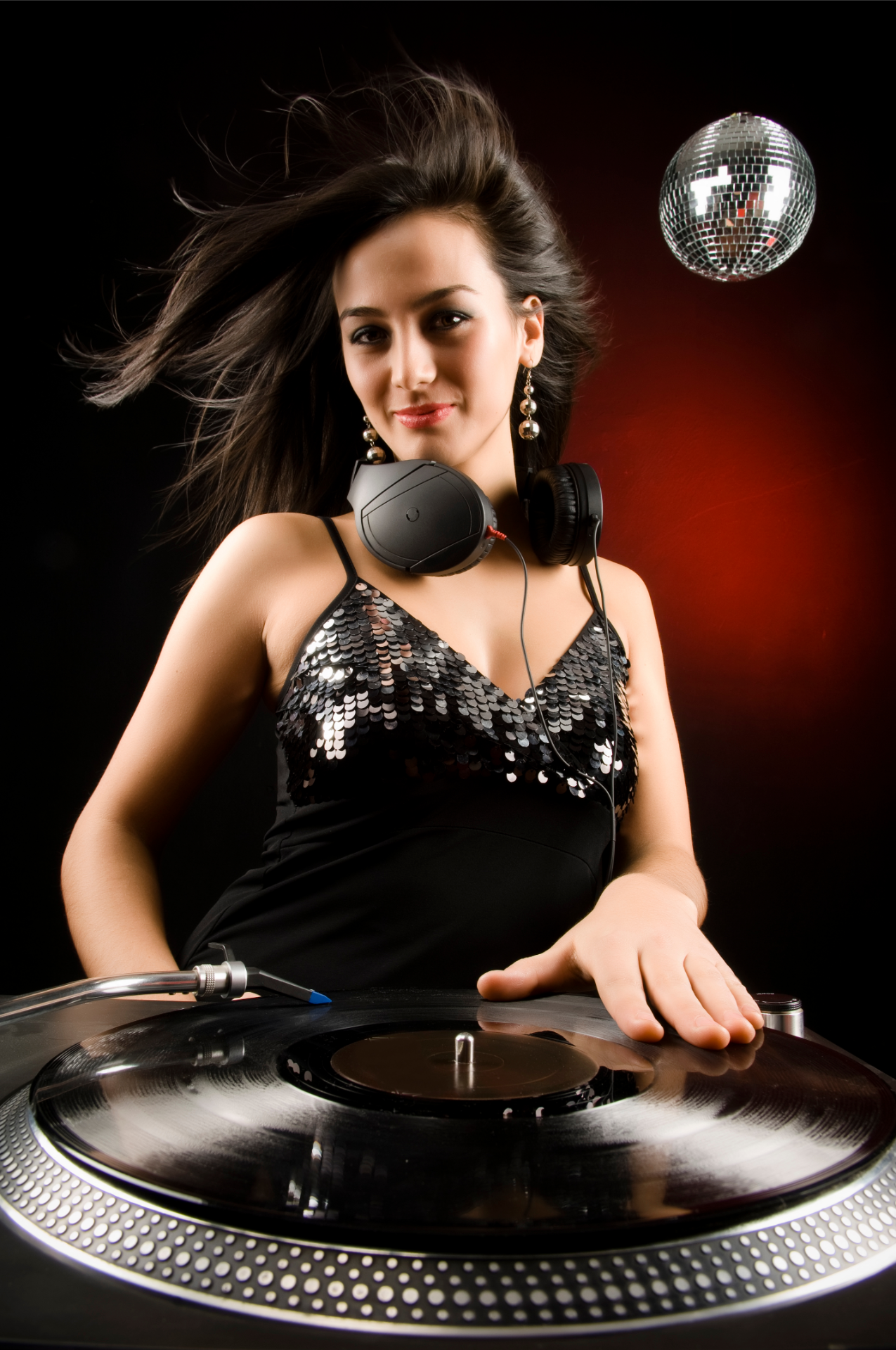 A person posing as a DJ at a turntable, wearing a sequined top and headphones, with a disco ball against a dark backdrop.