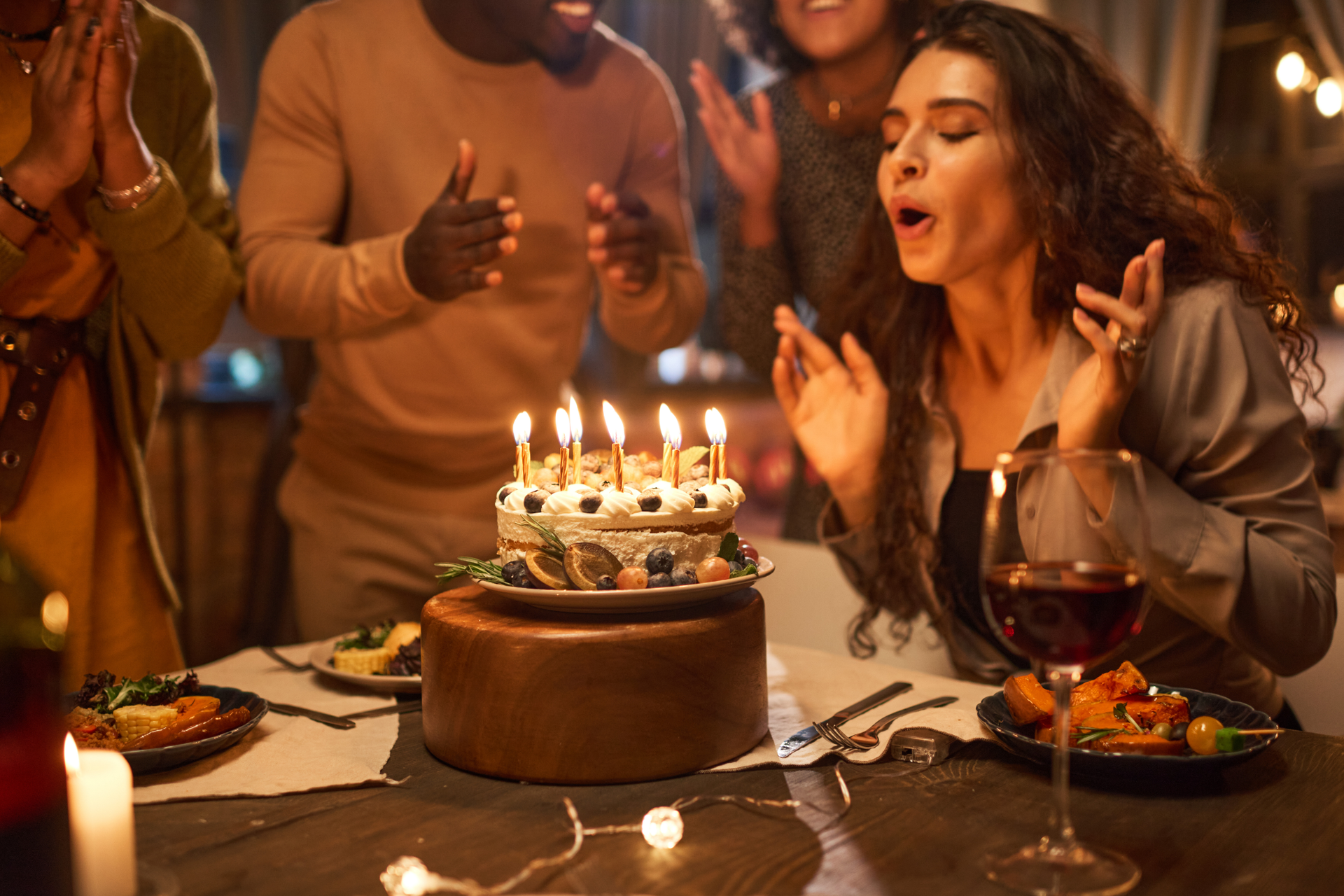 A person blows out candles on a birthday cake at a table surrounded by friends in a dimly lit setting.