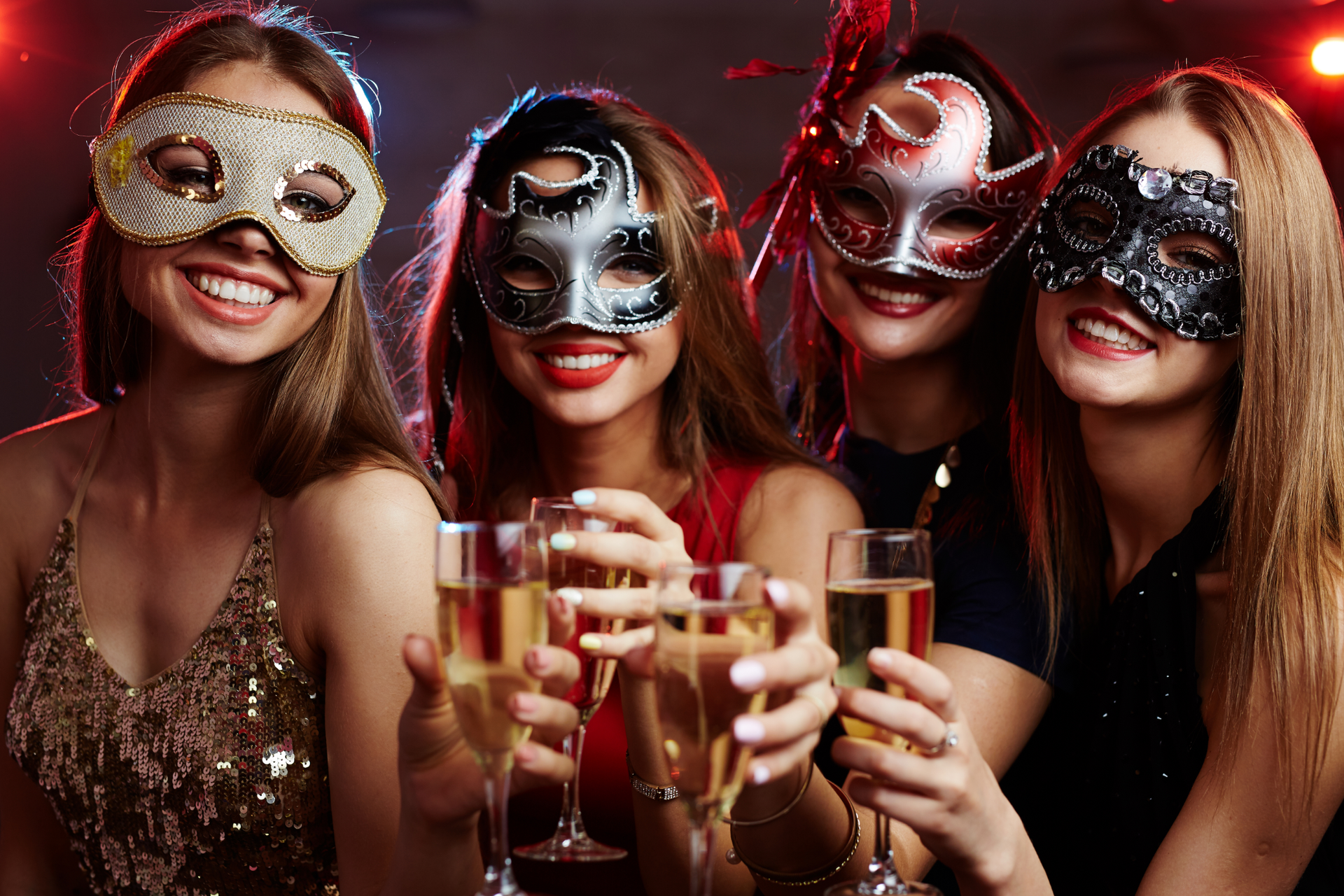 Four smiling people in masquerade masks holding champagne flutes and toasting together in a dimly lit, festive setting.