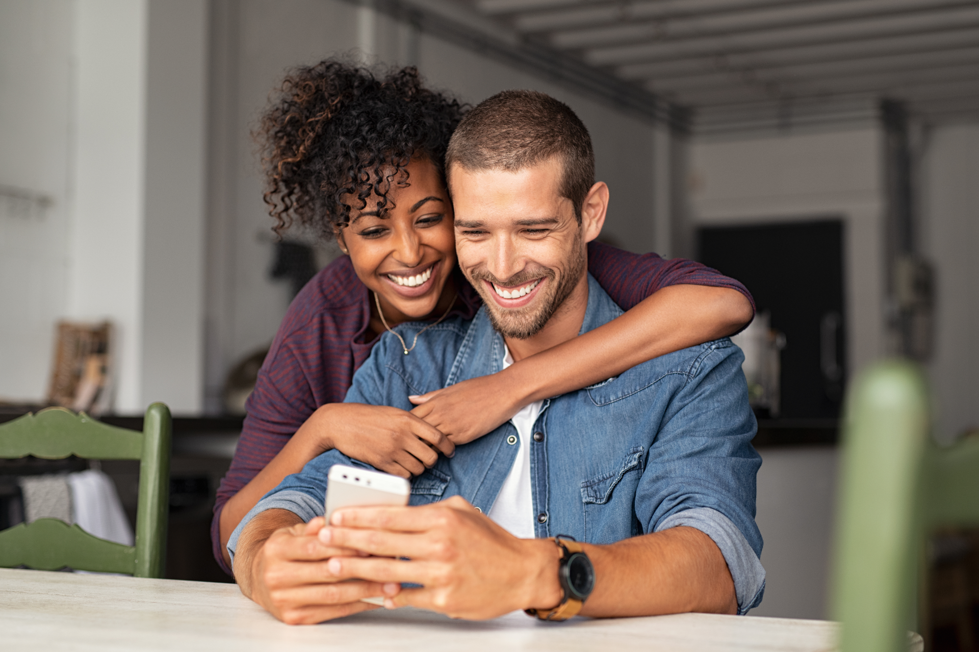 A smiling person hugging another from behind as they both look at a smartphone held in their hands.