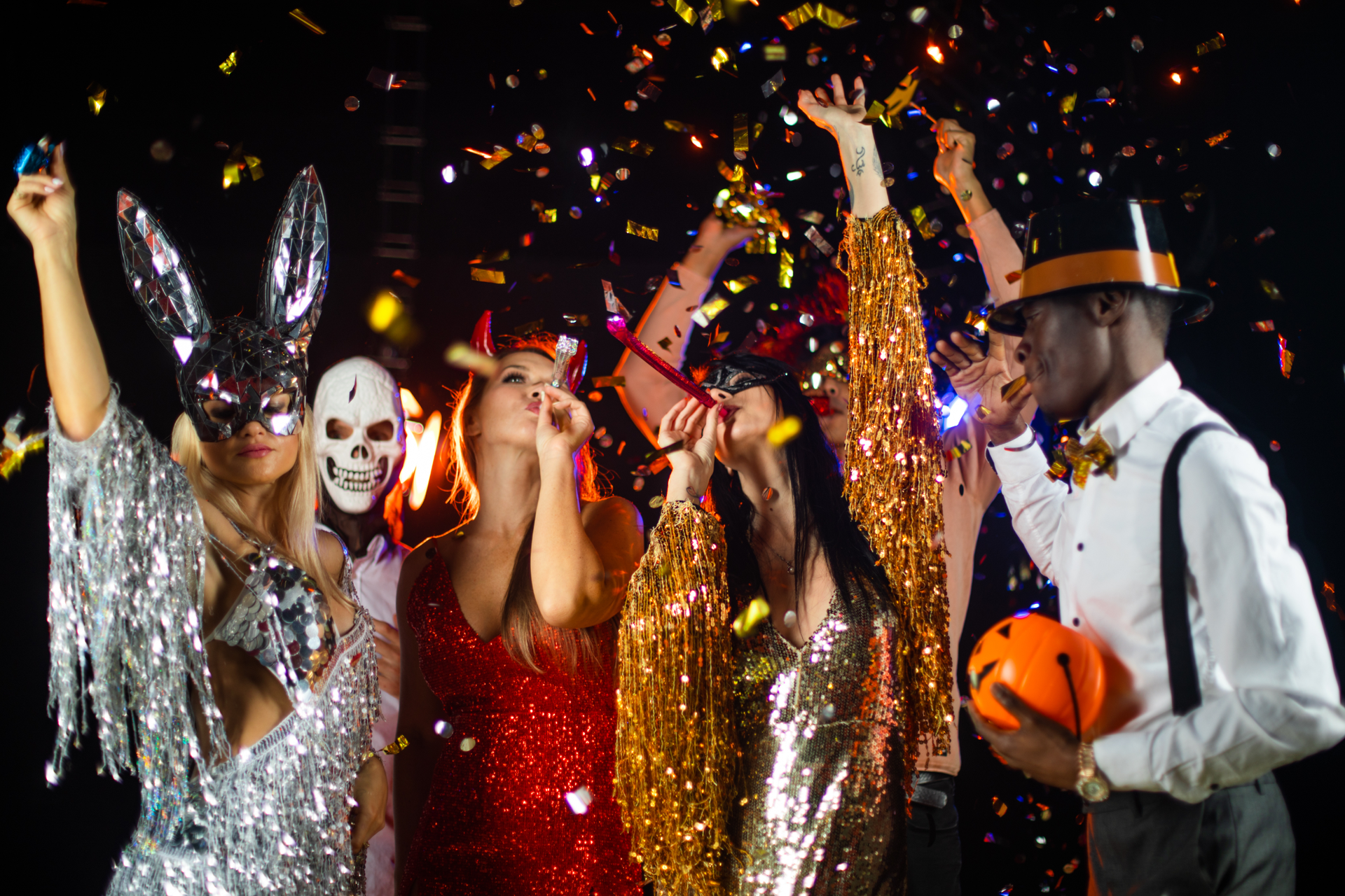 A group of people in costumes and masks celebrate at a party with gold confetti falling against a dark background.