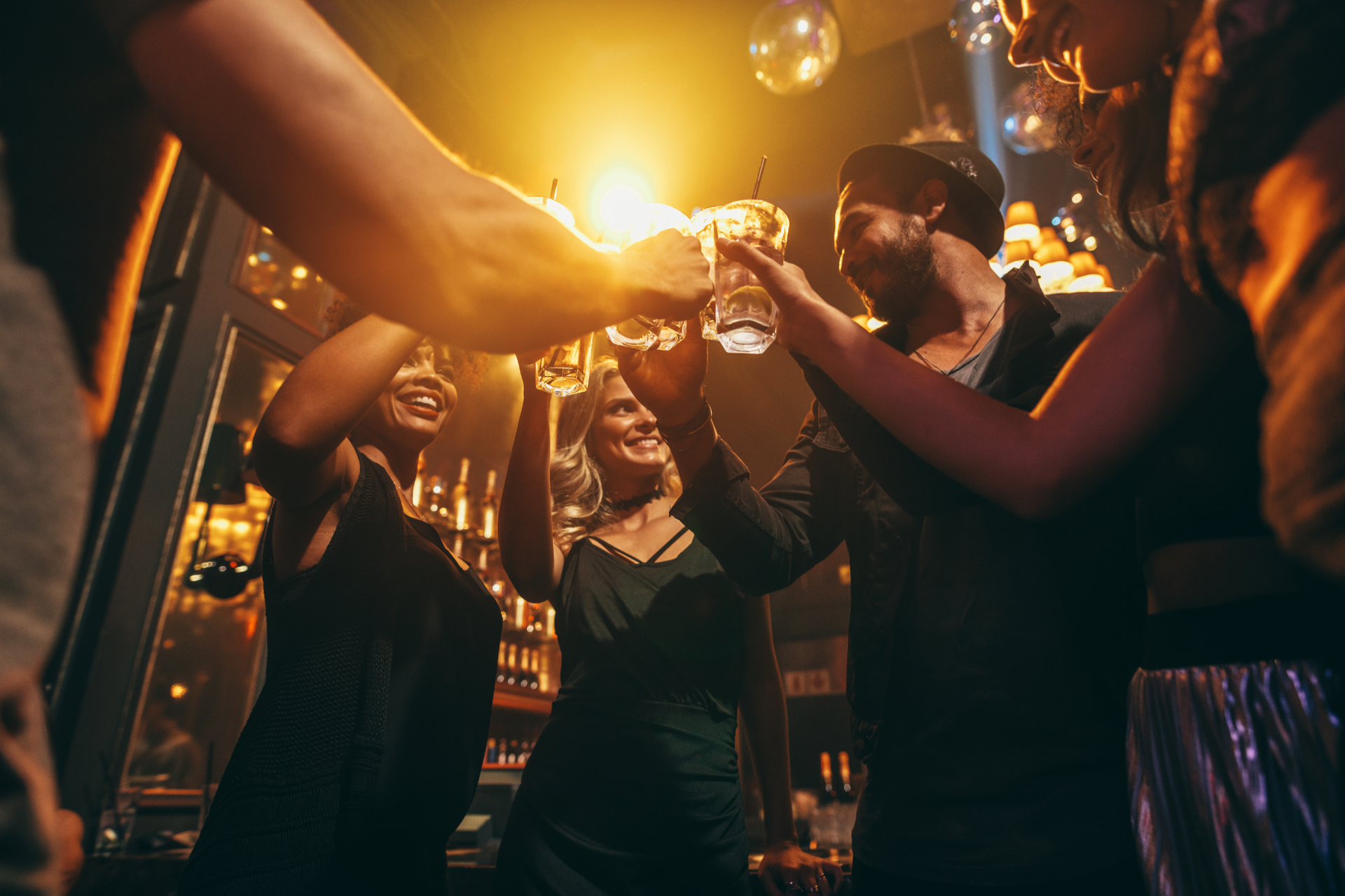 A group of friends in a dimly lit bar toast with cocktails, laughing under warm golden light.