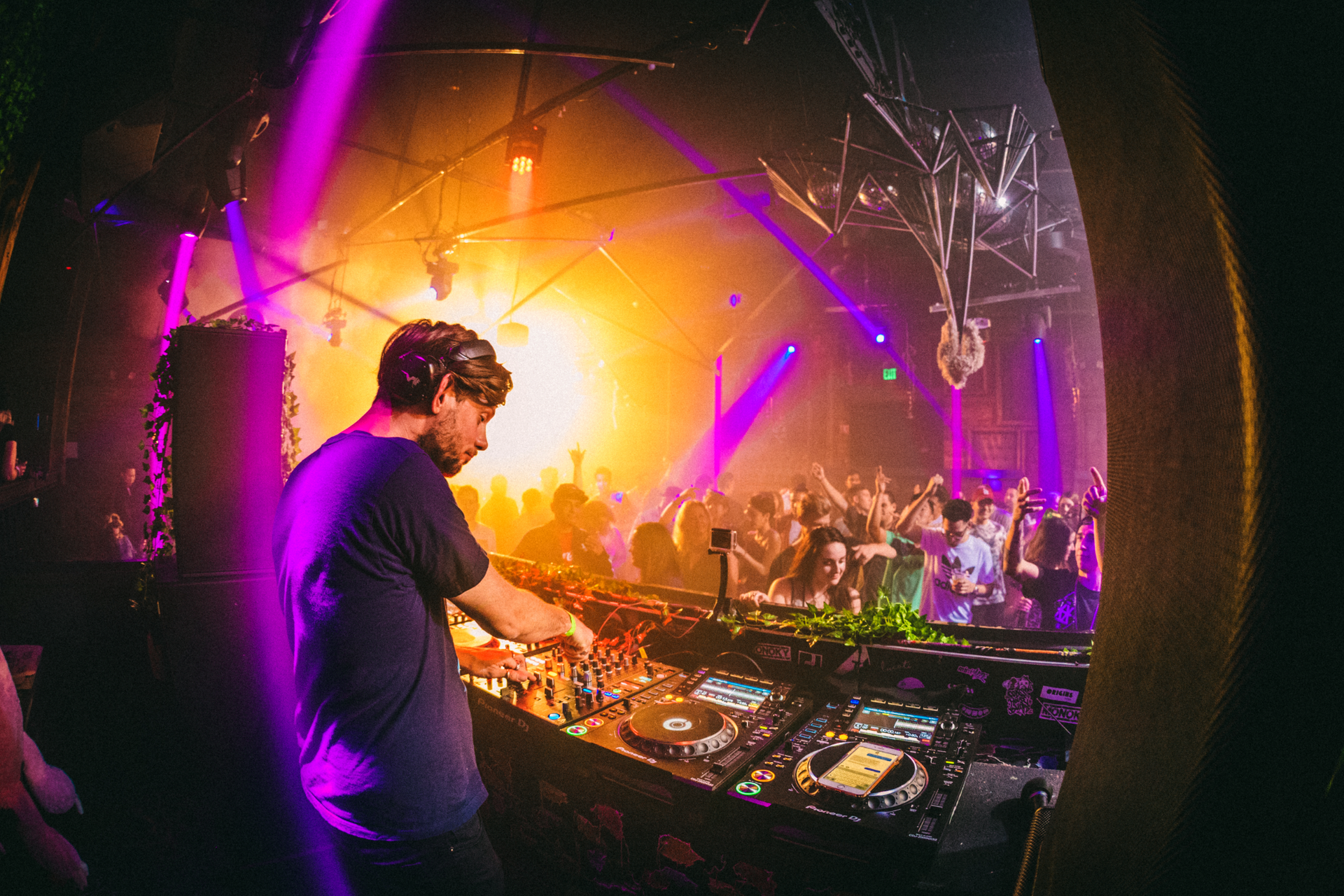 A DJ performs at a lively nightclub with vibrant purple and orange stage lights, blurred crowds, and mixing equipment.