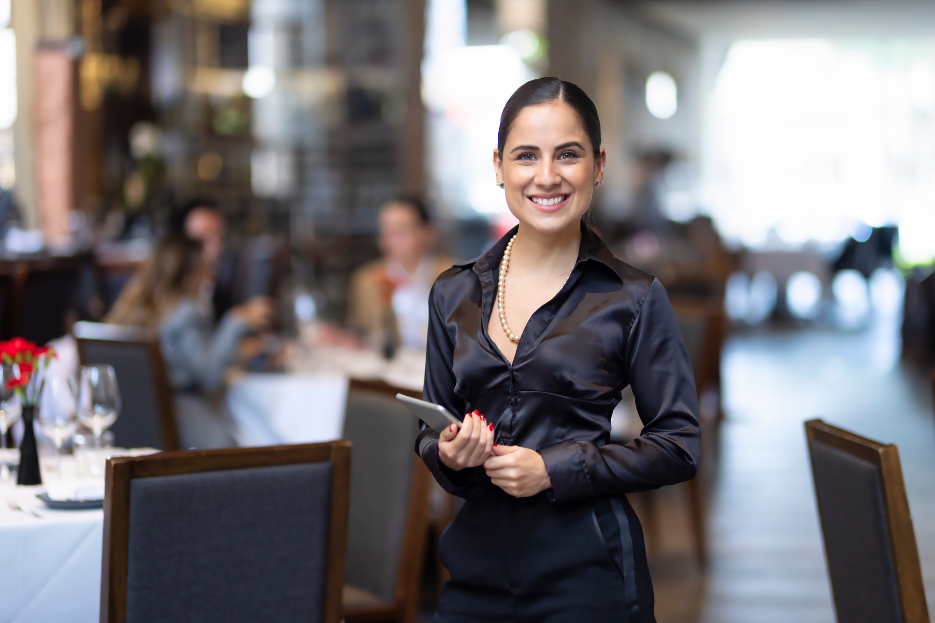 A smiling server in a black uniform stands in a restaurant, holding a digital device with customers in the background.