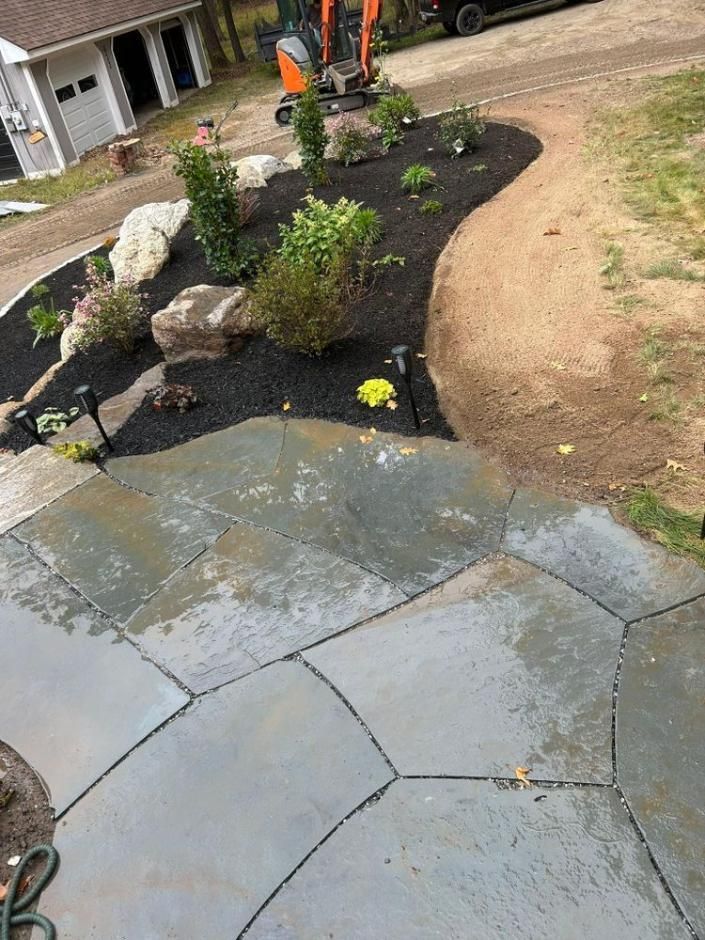 Garden bed with black mulch, rocks, and plants beside a wet gray patio and dirt path