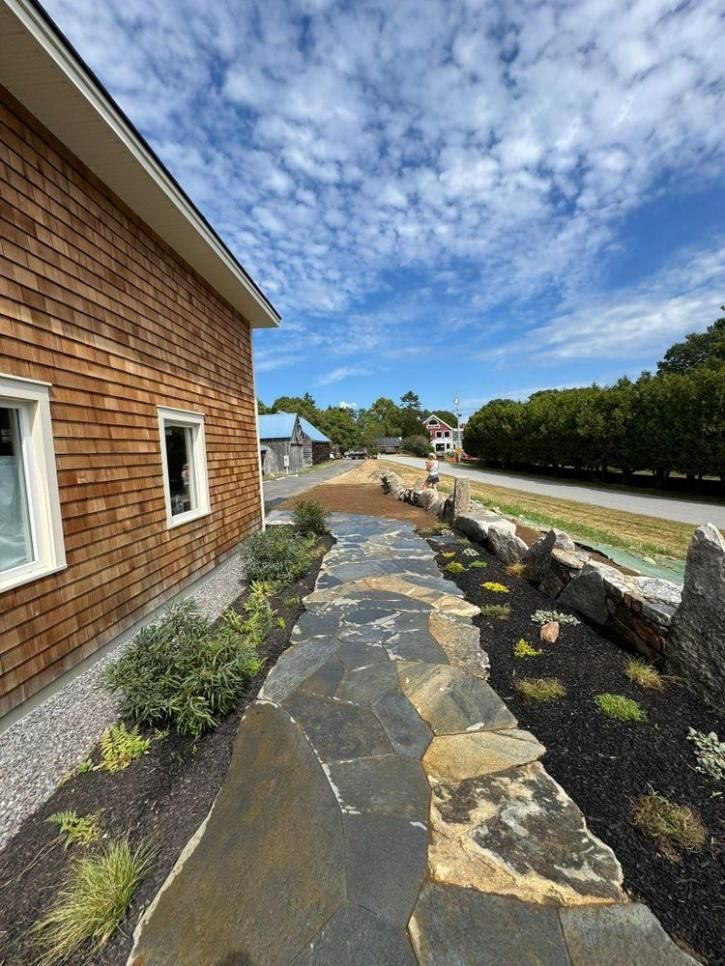 Stone walkway beside a brick house, with landscaped beds and a blue sky above.
