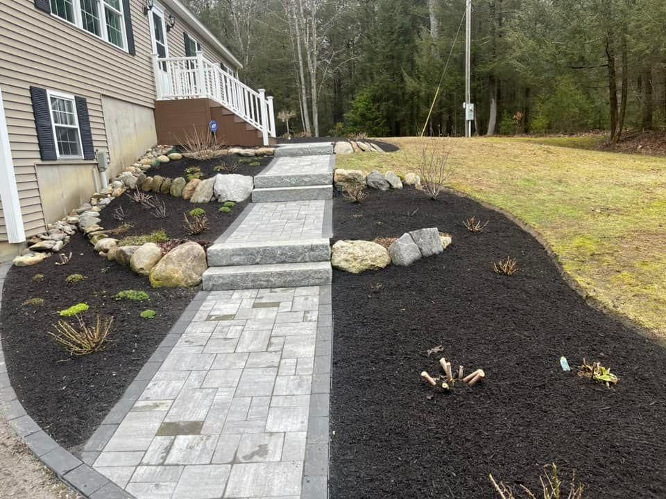 Stone walkway and steps with black mulch landscaping beside a house in a wooded yard.