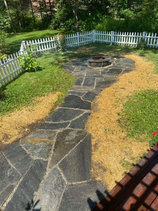 Backyard stone path leading to a fire pit, bordered by dry grass and a white picket fence.