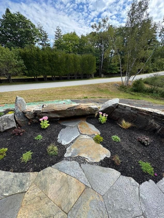 Rock garden with flagstone path, stone bench, and small flowers beside a tree-lined road