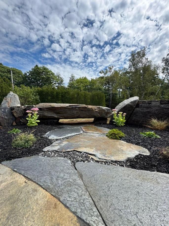 Stone garden pathway with a rustic bench, rocks, flowers, and trees under a cloudy blue sky
