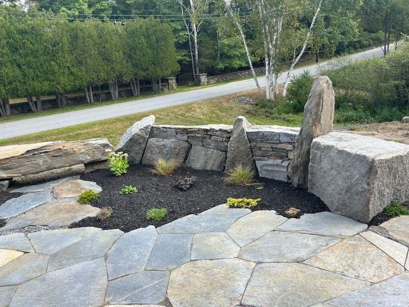 Stone garden with large rocks, dark mulch, and a road and trees in the background