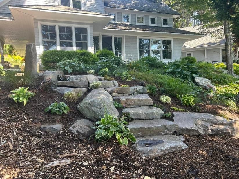Stone steps and boulders leading to a house with a landscaped front yard and shrubs.