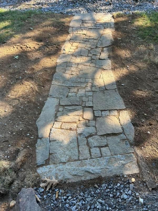 Stone walkway path bordered by dirt and grass, with dappled sunlight on the stones