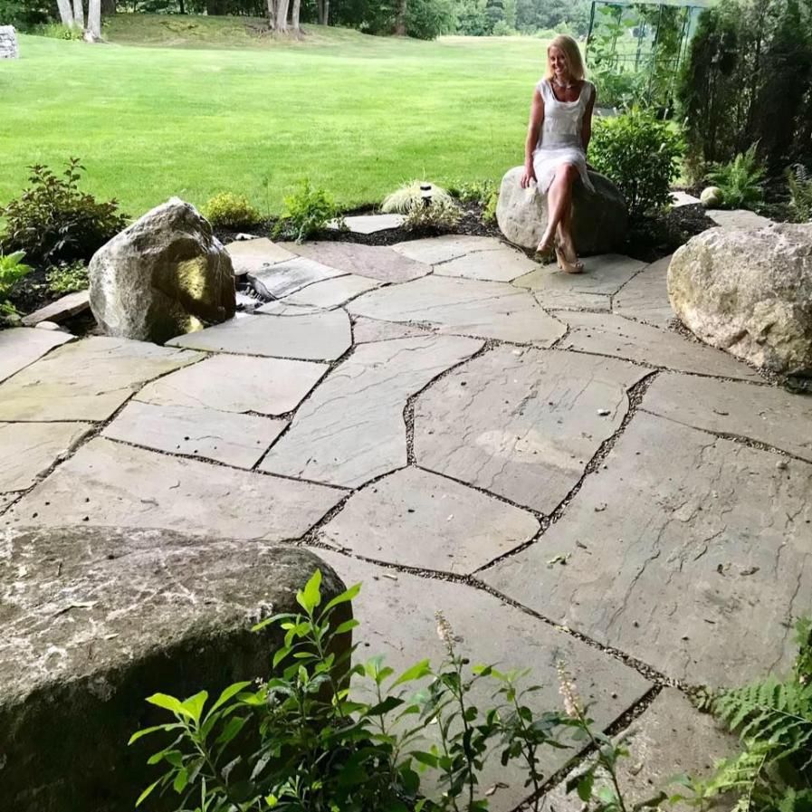 Person sitting on a stone patio beside a grassy garden with rocks and trees