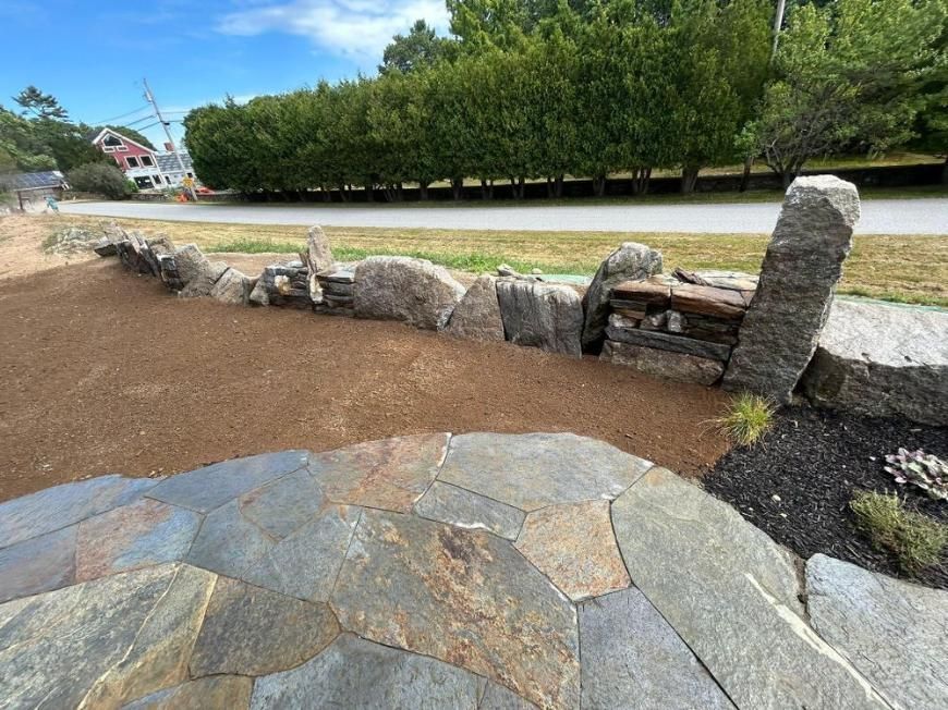 Stone walkway beside a low rock wall and trees under a blue sky in a park-like setting