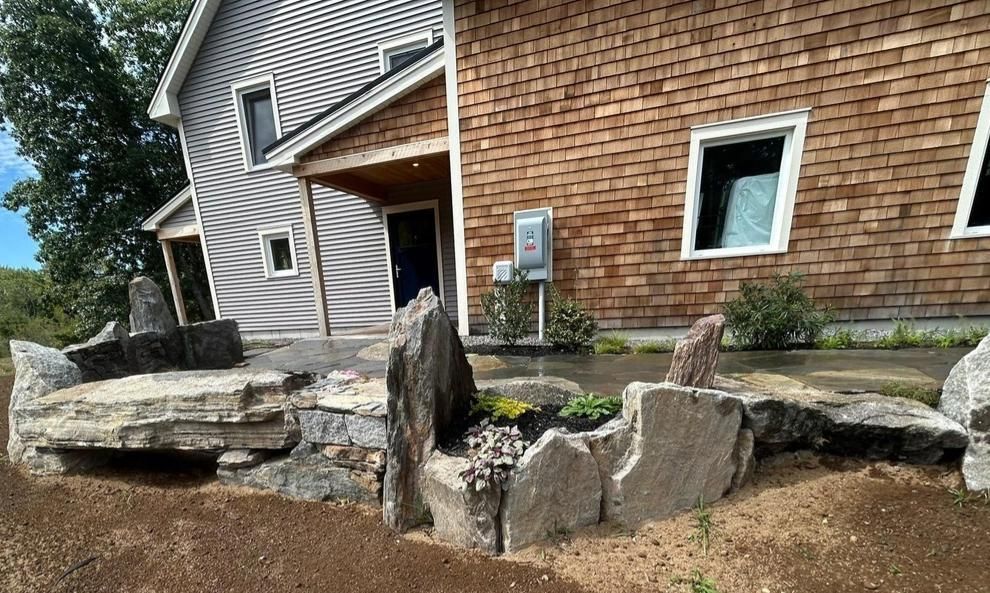 Rock retaining wall and walkway beside a house with brown shingles and a striped awning