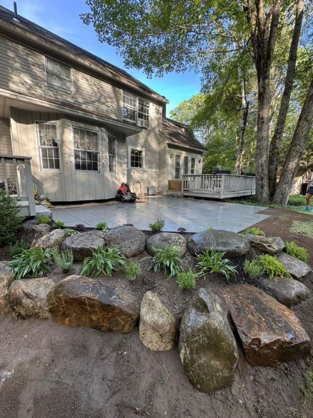 Rustic house with stone terrace, flower bed, and trees under a blue sky; person sitting by the wall.