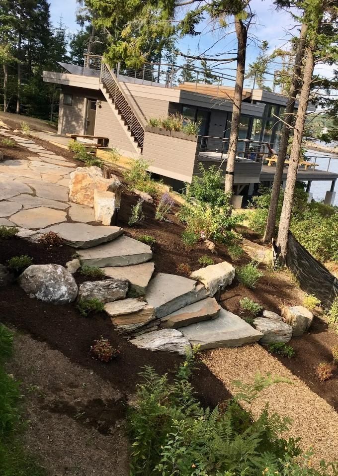 Modern house on a rocky hillside with stone steps, trees, and a view of the water