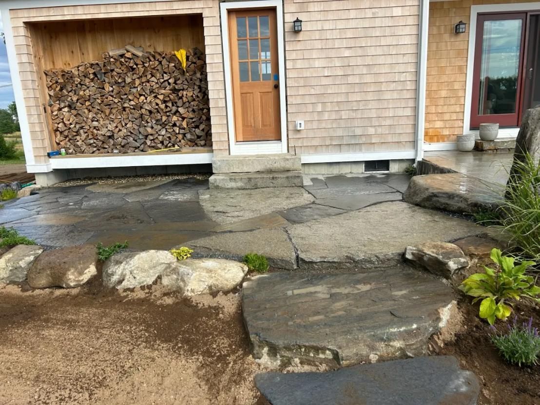 House entry with stone steps, walkway, and a woodpile stacked in an open garage bay