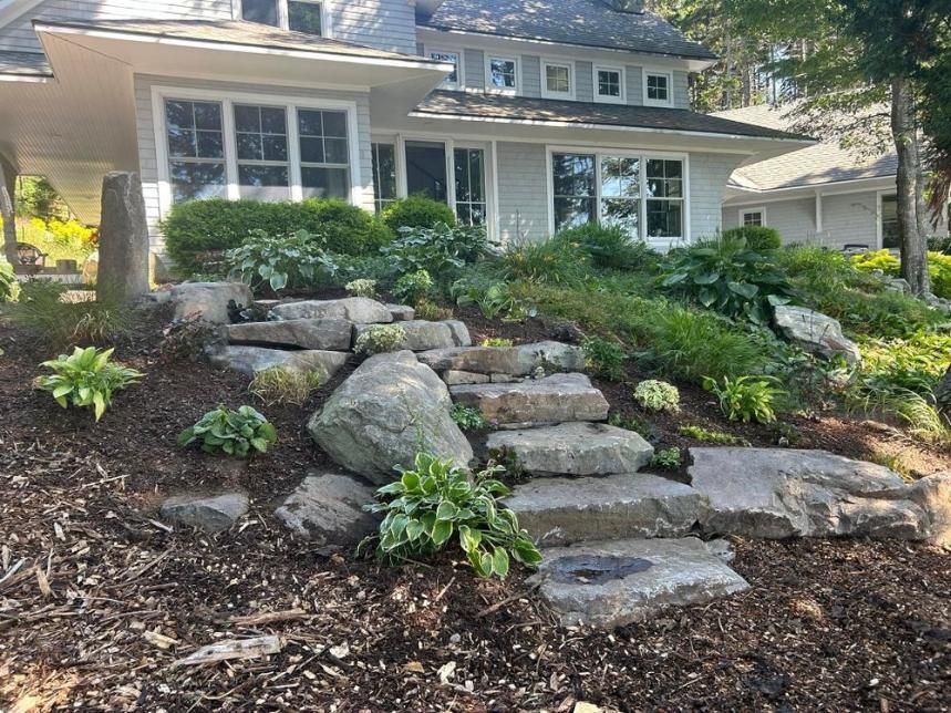 Front yard stone steps and boulders leading to a house with white trim and landscaped shrubs
