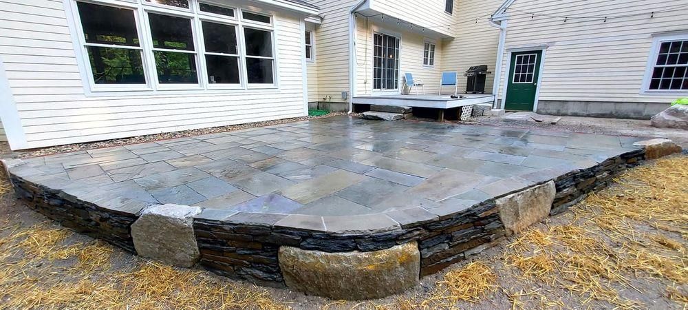Stone patio beside a white house with a wooden deck and green door in a yard.