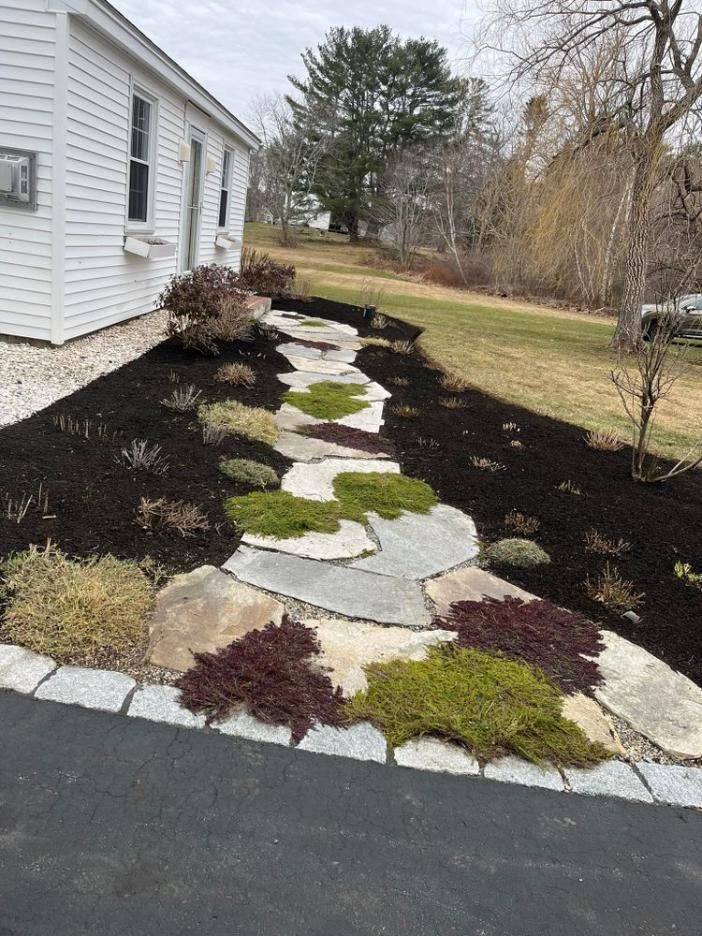 Landscaped front walkway with stepping stones, moss, and mulch beside a white house