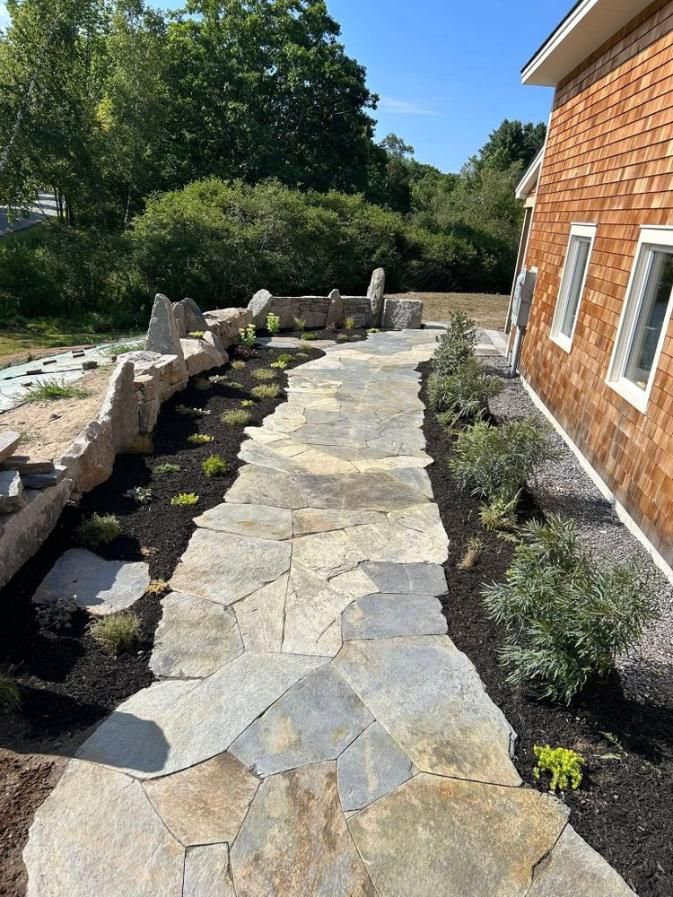 Stone garden path beside a brick house, bordered by rocks and low shrubs in a sunny yard