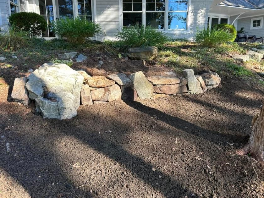 Stone retaining wall in front of a house, with shrubs and a dirt yard in the foreground