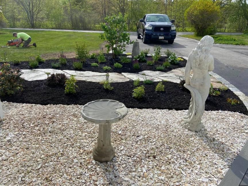 Garden with black mulch, small shrubs, a birdbath, and a white statue beside a road; car in background
