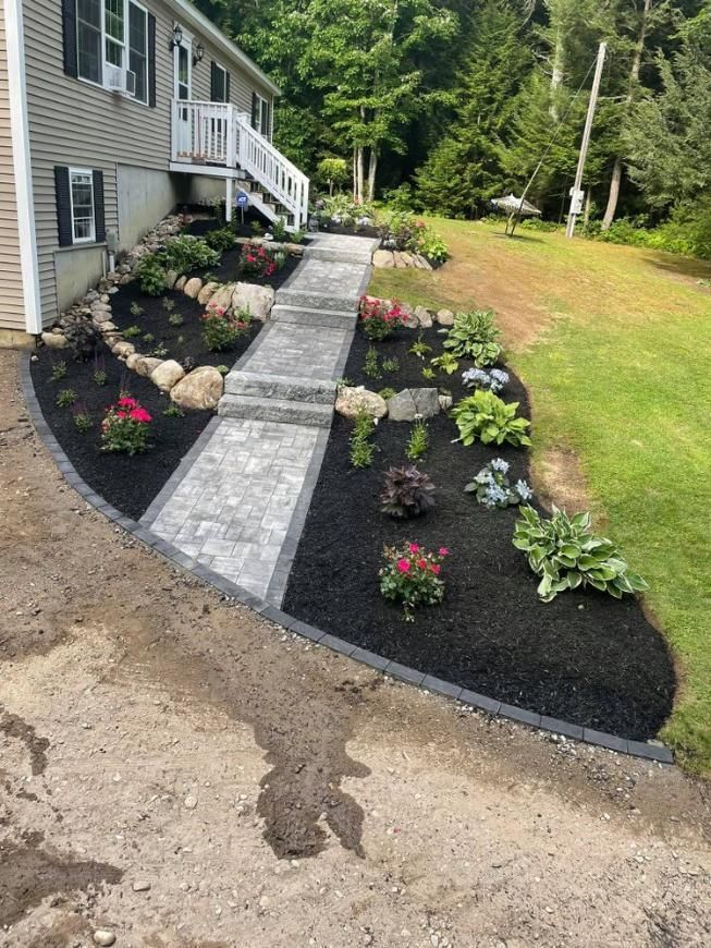 Stone walkway through black mulch flower beds beside a house, with pink and white flowers and green lawn.