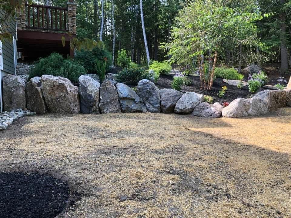 Rocky backyard garden with a wooden deck, trees, and a dirt clearing near a stone border
