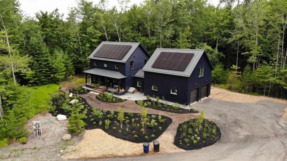 Aerial view of two dark houses with solar panels, surrounded by trees, a landscaped yard, and gravel driveway