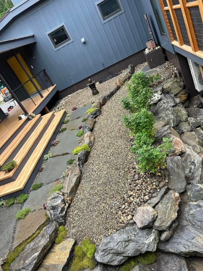 Narrow gravel and stone garden bed beside a gray house with small green plants and stepping stones