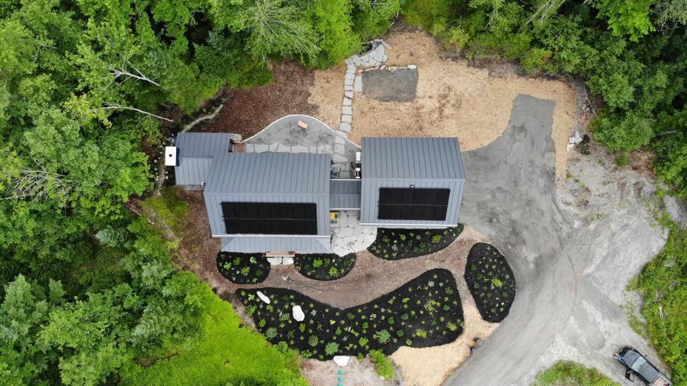 Aerial view of a modern gray house with black landscaping, surrounded by dense green trees and a gravel driveway