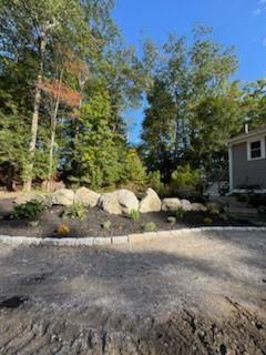 Rock garden and trees beside a gravel driveway next to a gray house under a blue sky