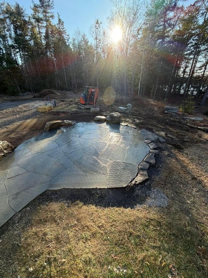 Sunlit backyard with a cracked concrete slab, trees, and a small excavator in the background.