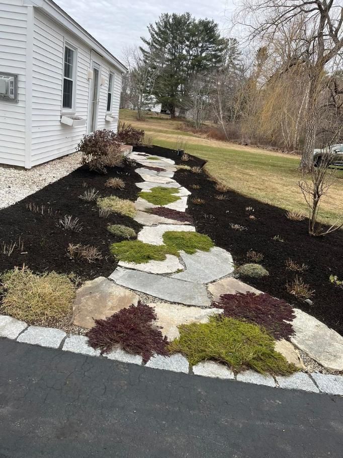 Curved stone walkway bordered by mulch and colorful low shrubs beside a white house.