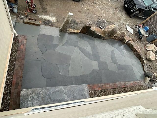 Newly paved gray patio beside a house, bordered by dirt and scattered stones, viewed from above