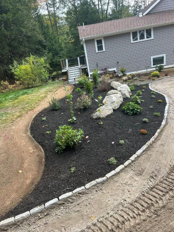 Curved garden bed with black mulch, white stones, shrubs, and a house beside a dirt driveway